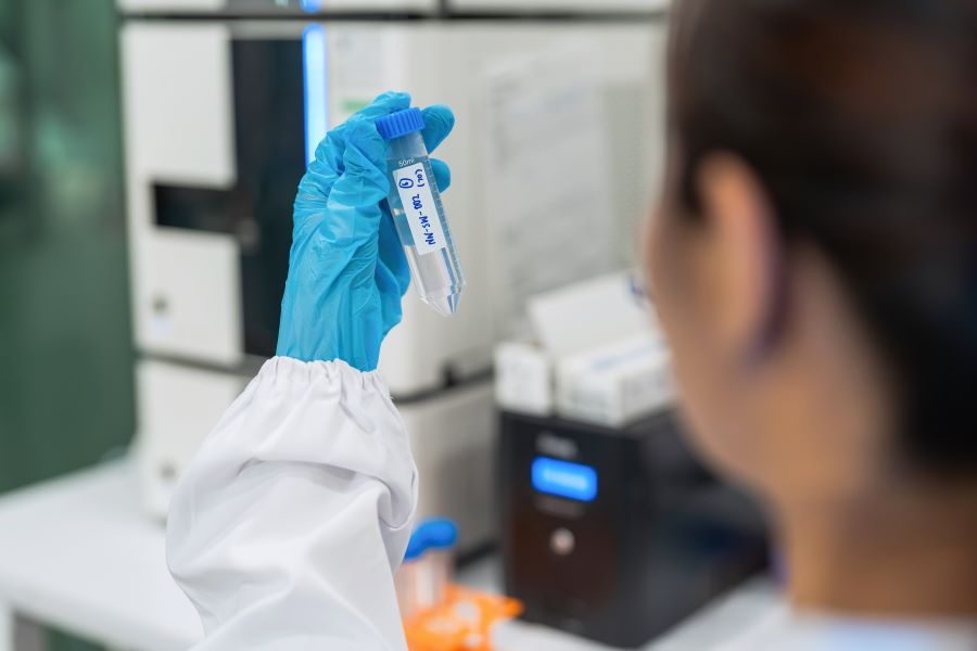 Lab technician in blue gloves holds a small sterile sample tube labeled with a blue handwritten tag in a clinical lab.